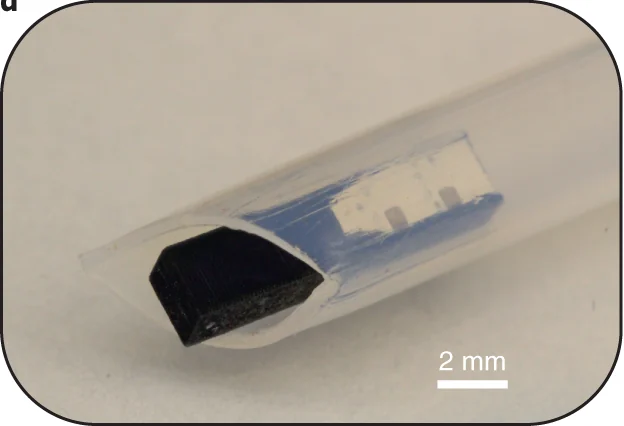 A small transparent ME-BIT capsule with a dark magnetoelectric strip and a tiny circuit visible inside, shown in a clear sheath.