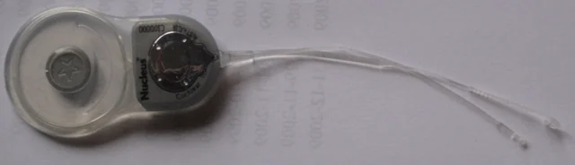 An archival Cochlear Nucleus Freedom CI24RE implant on paper, with a round receiver coil, metal stimulator housing and two thin electrode leads.