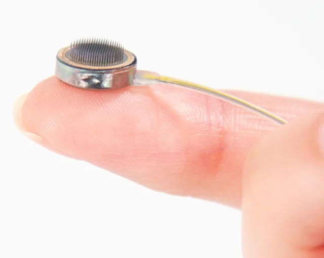 A Connexus cortical module resting on a fingertip, with a circular metal enclosure and a dense bed of fine penetrating electrodes.